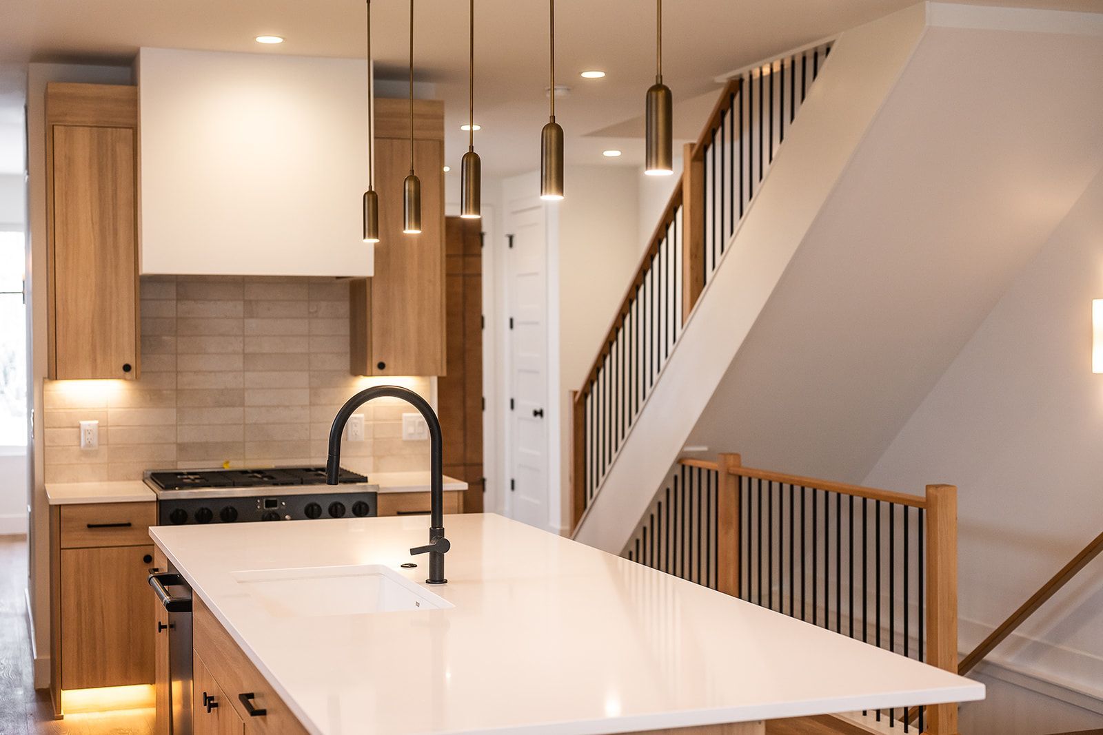 Modern kitchen with light wood cabinets, white countertops, and black fixtures; staircase in background.