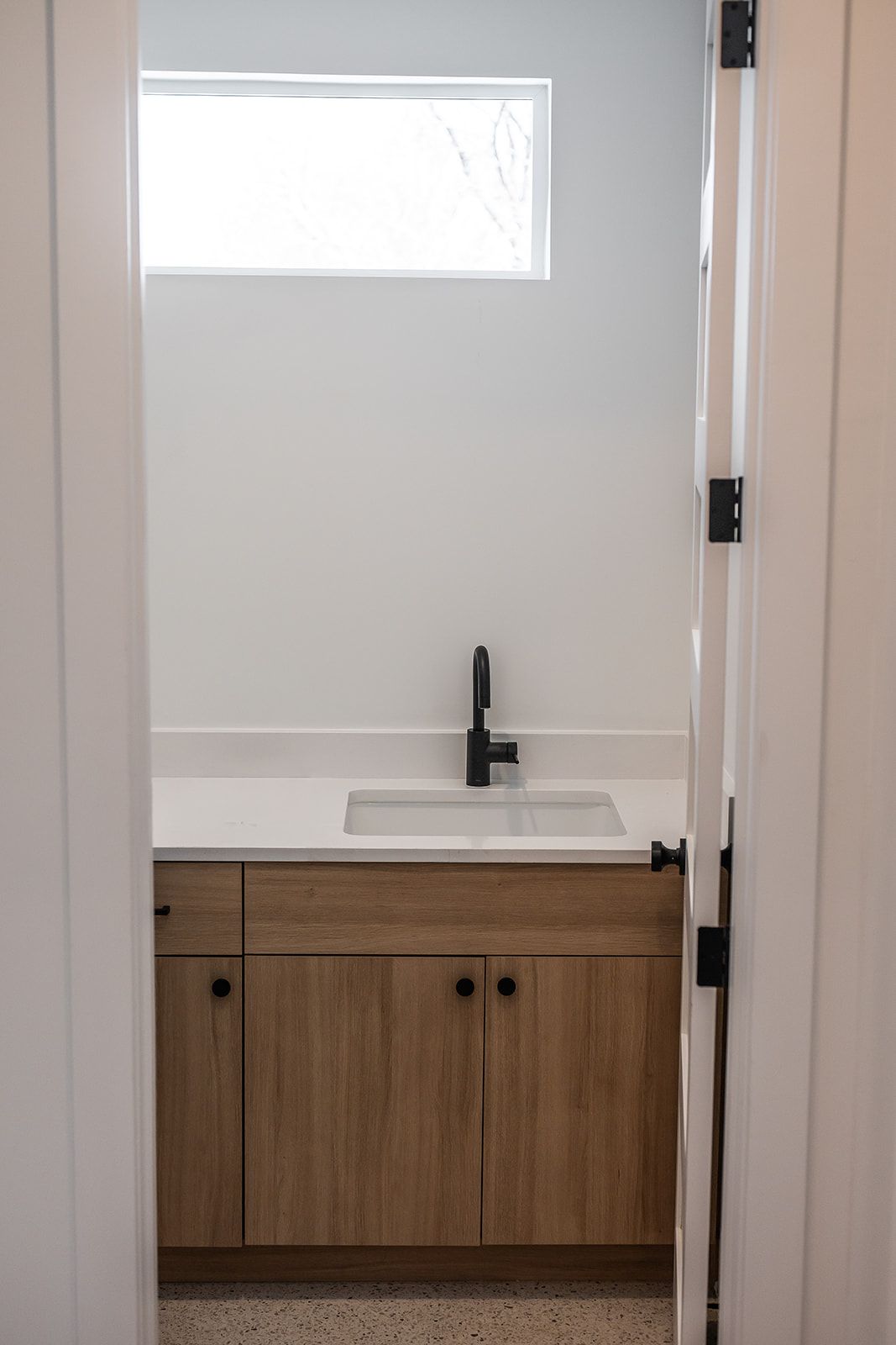 Bathroom with wood cabinets, white countertop and walls, black faucet, and rectangular window.