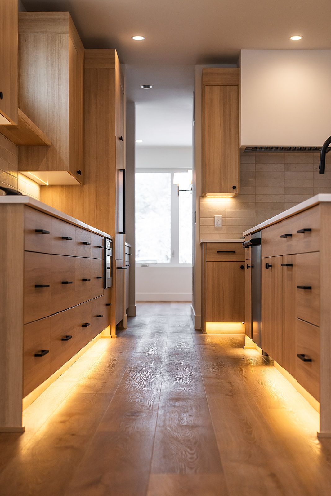 Modern kitchen with light wood cabinets, under-cabinet lighting, and a view to a window.