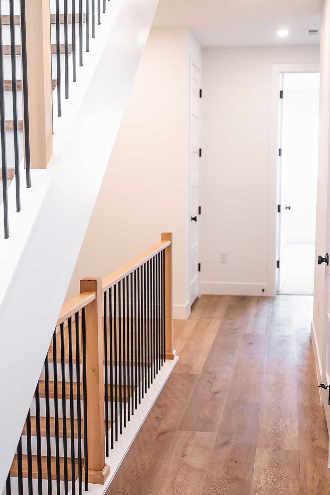 Hallway with hardwood floor, stairs with black spindles and wood handrail, white walls, and black doors.