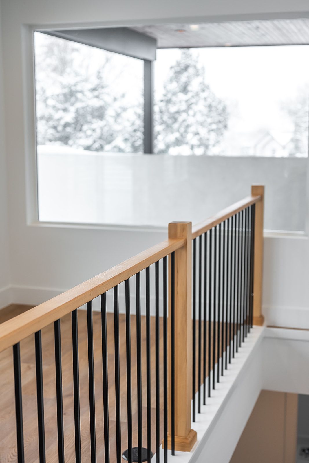 Wooden railing with black vertical bars near a window overlooking a snowy landscape.