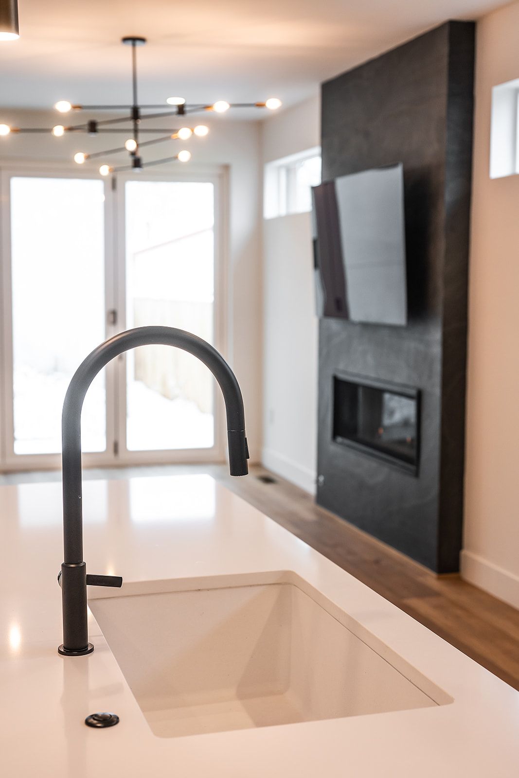 Black kitchen faucet over white sink on countertop. Modern kitchen with fireplace and glass doors.
