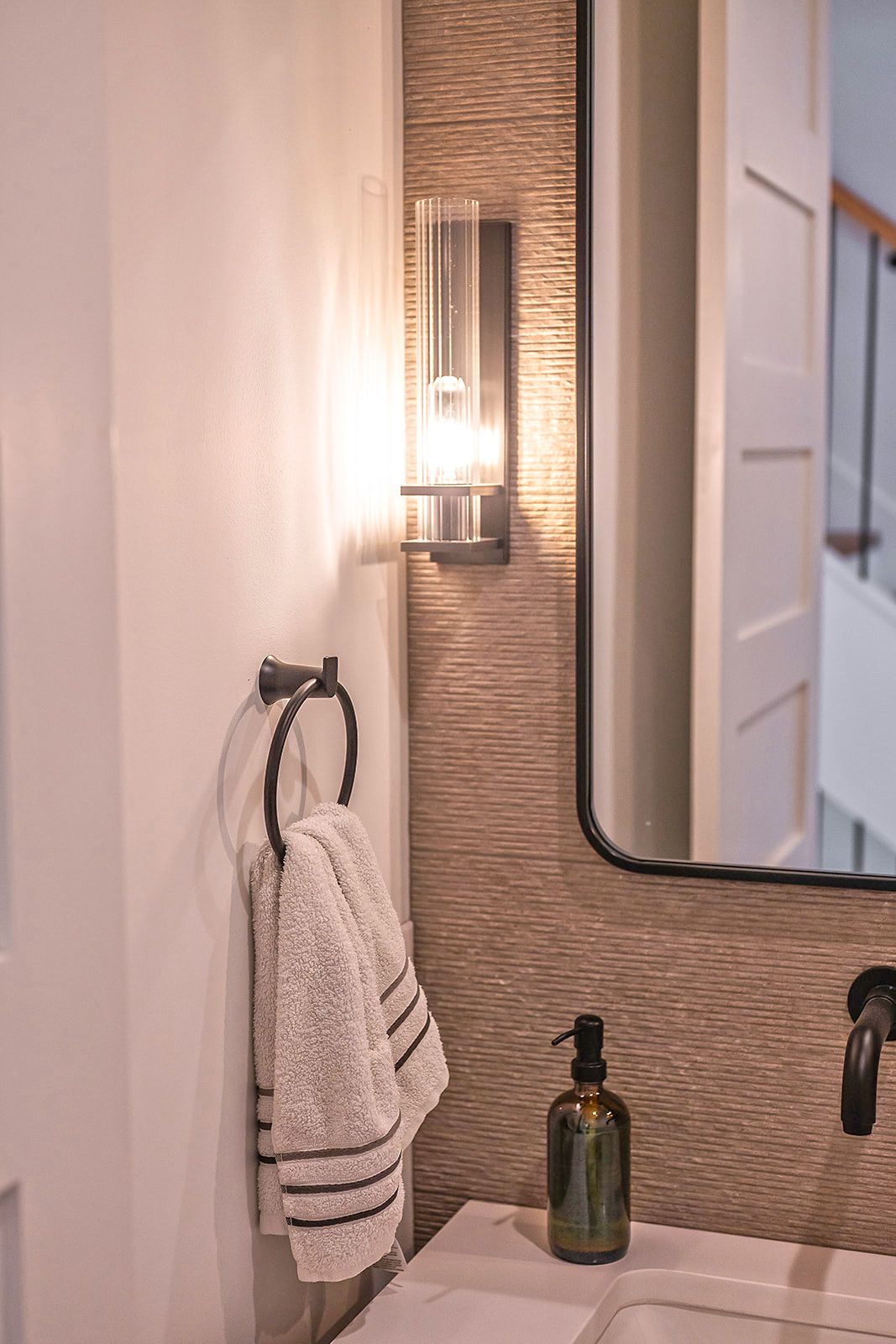 A bathroom with a wall-mounted light, towel ring, mirror, and soap dispenser. Textured stone wall.