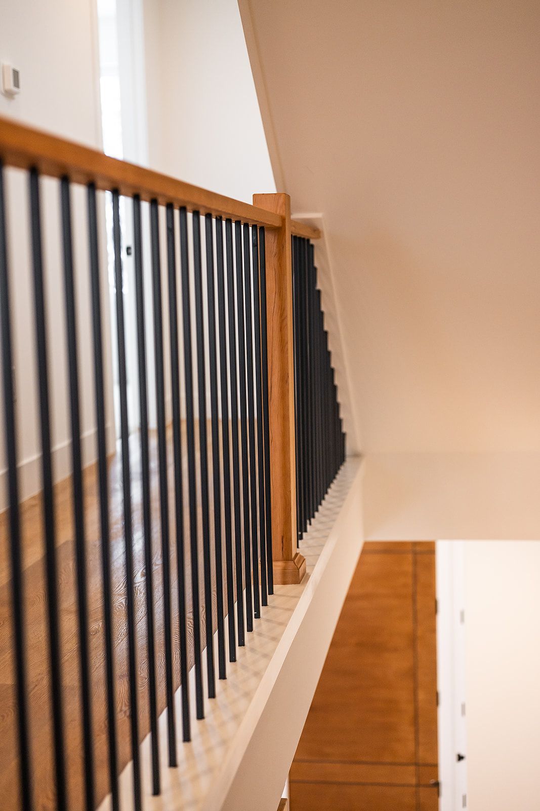 Wooden railing with black vertical bars along a second-floor hallway overlooking a lower level with a wooden door.