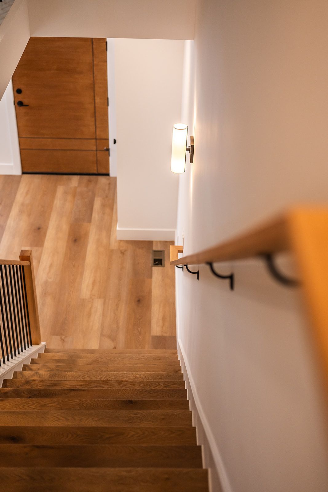 View down a staircase with wooden steps and railing, leading to a door and wood flooring.
