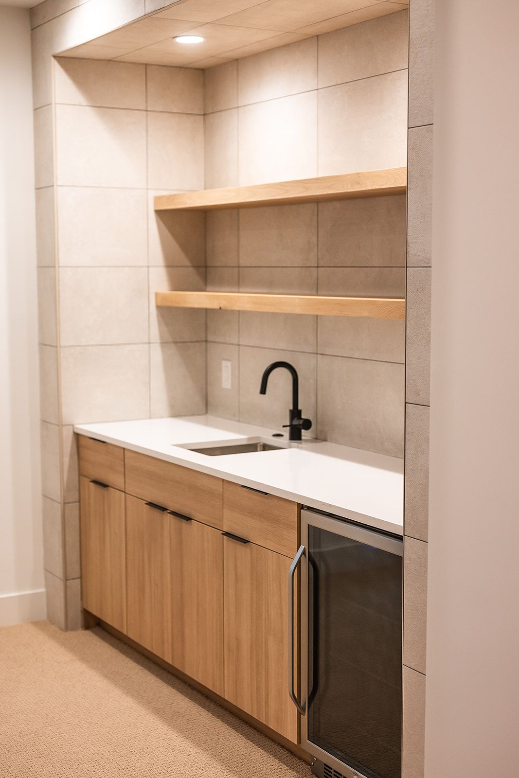 Modern wet bar with light wood cabinets, white countertops, black faucet, and built-in wine fridge.
