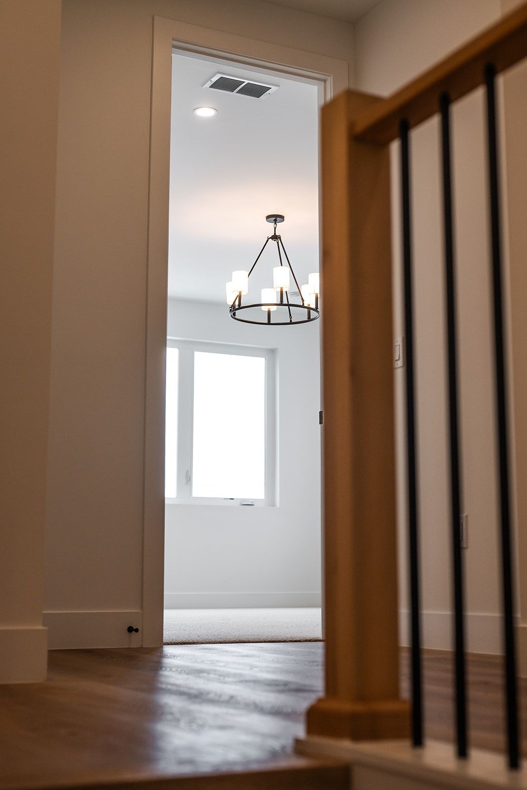 Hallway view through a doorway with a chandelier and window at the end. Wooden staircase on the right.