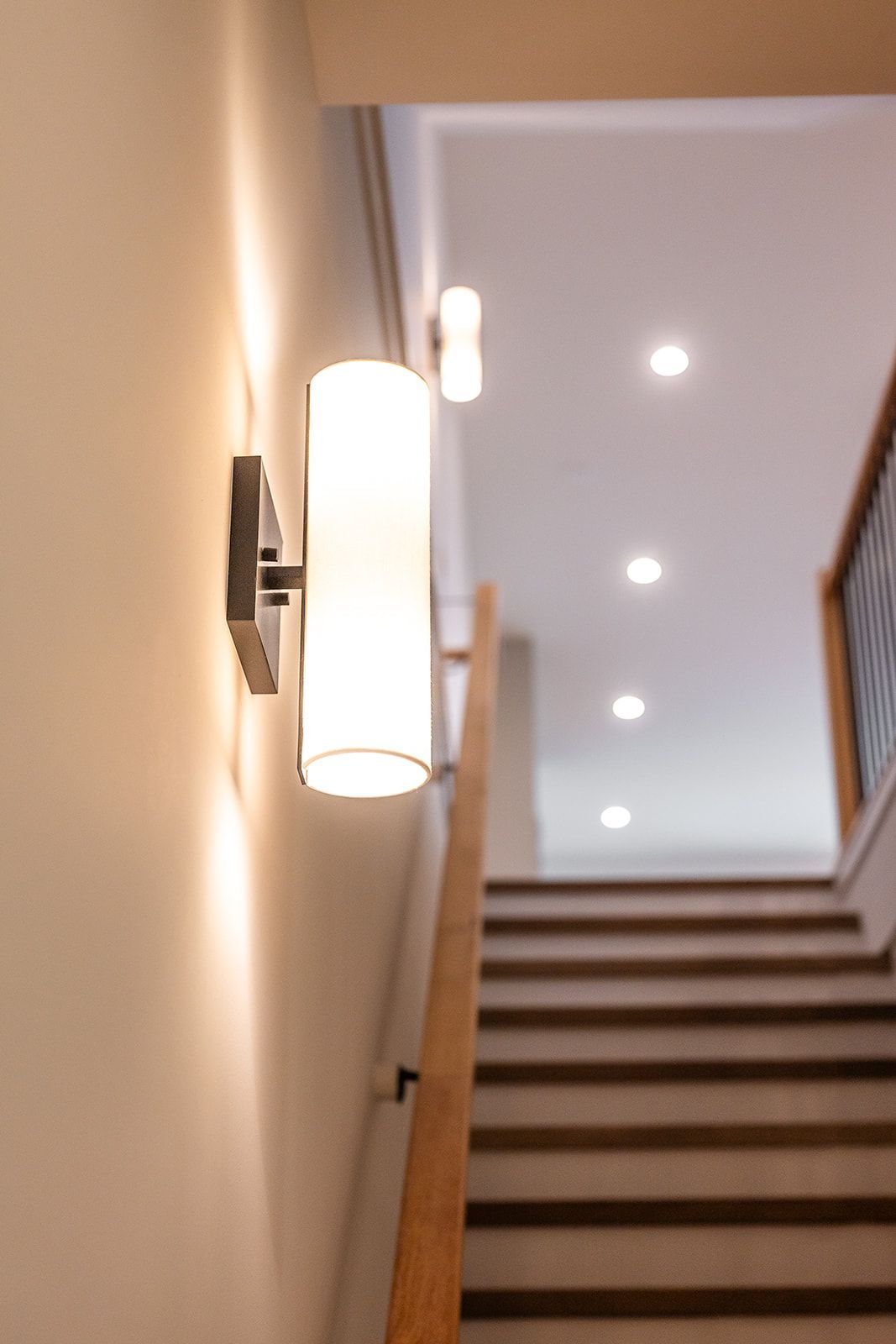 Wall-mounted cylindrical light fixture illuminates a stairwell. Stairs lead upwards to recessed ceiling lights.