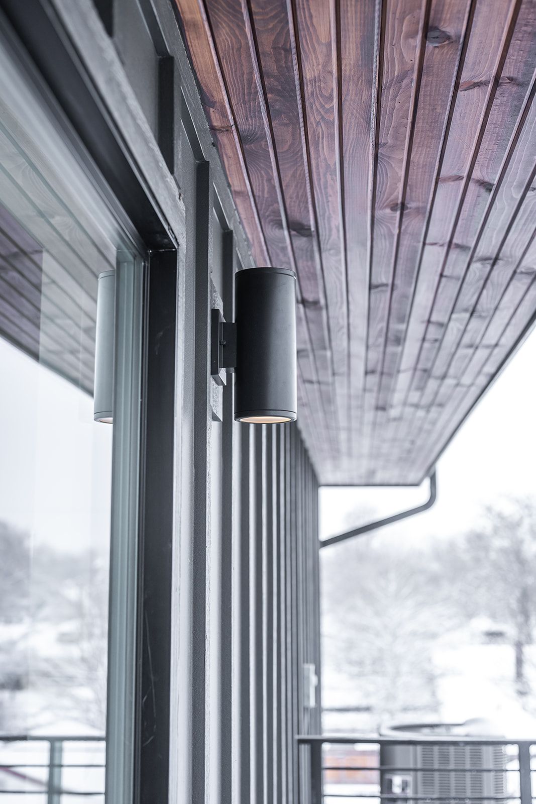 Black cylindrical outdoor lights on a building's exterior, with wooden ceiling and snowy background.