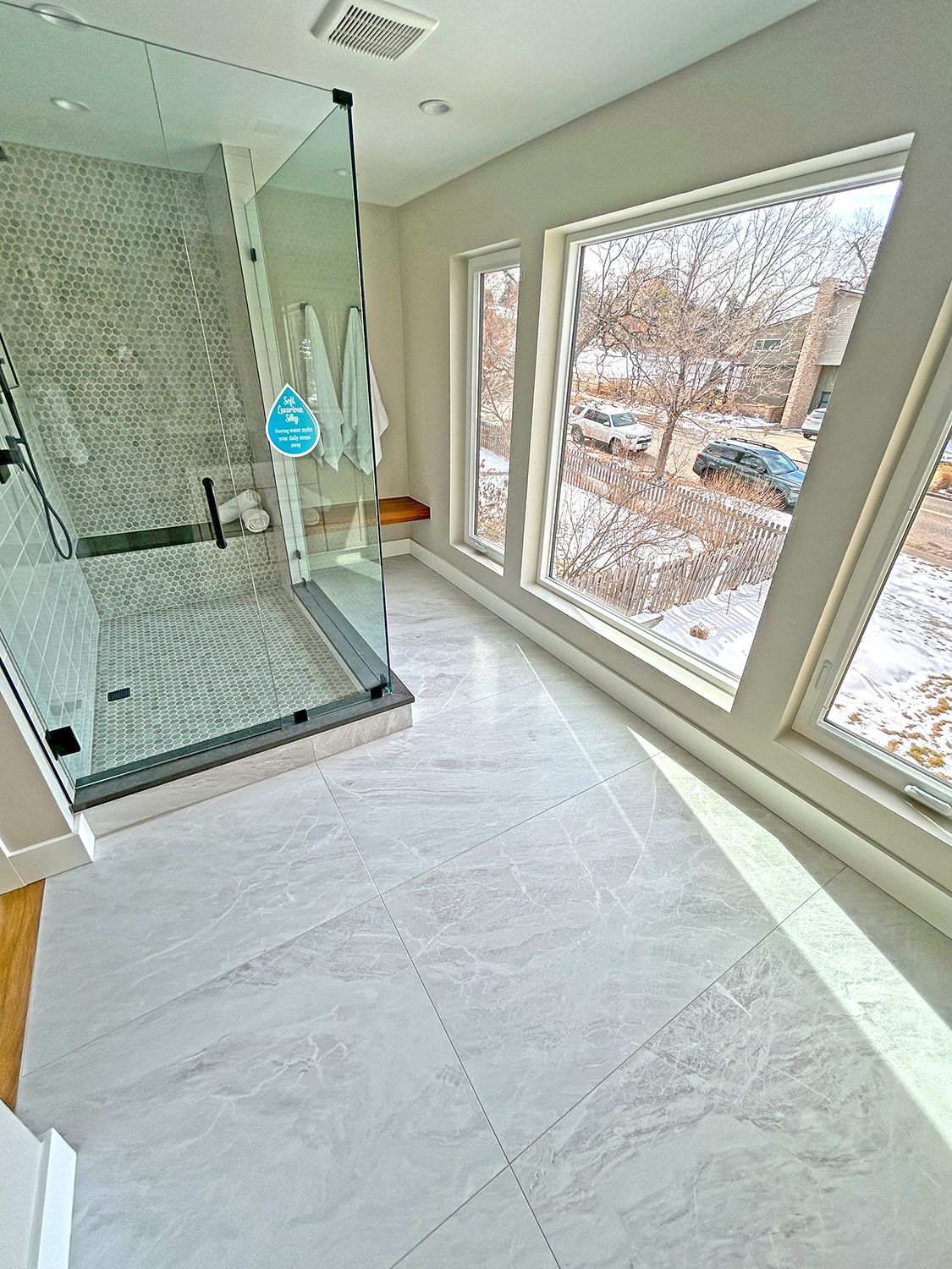 Spacious bathroom with glass shower, light gray tile floor, and large windows with outdoor view.