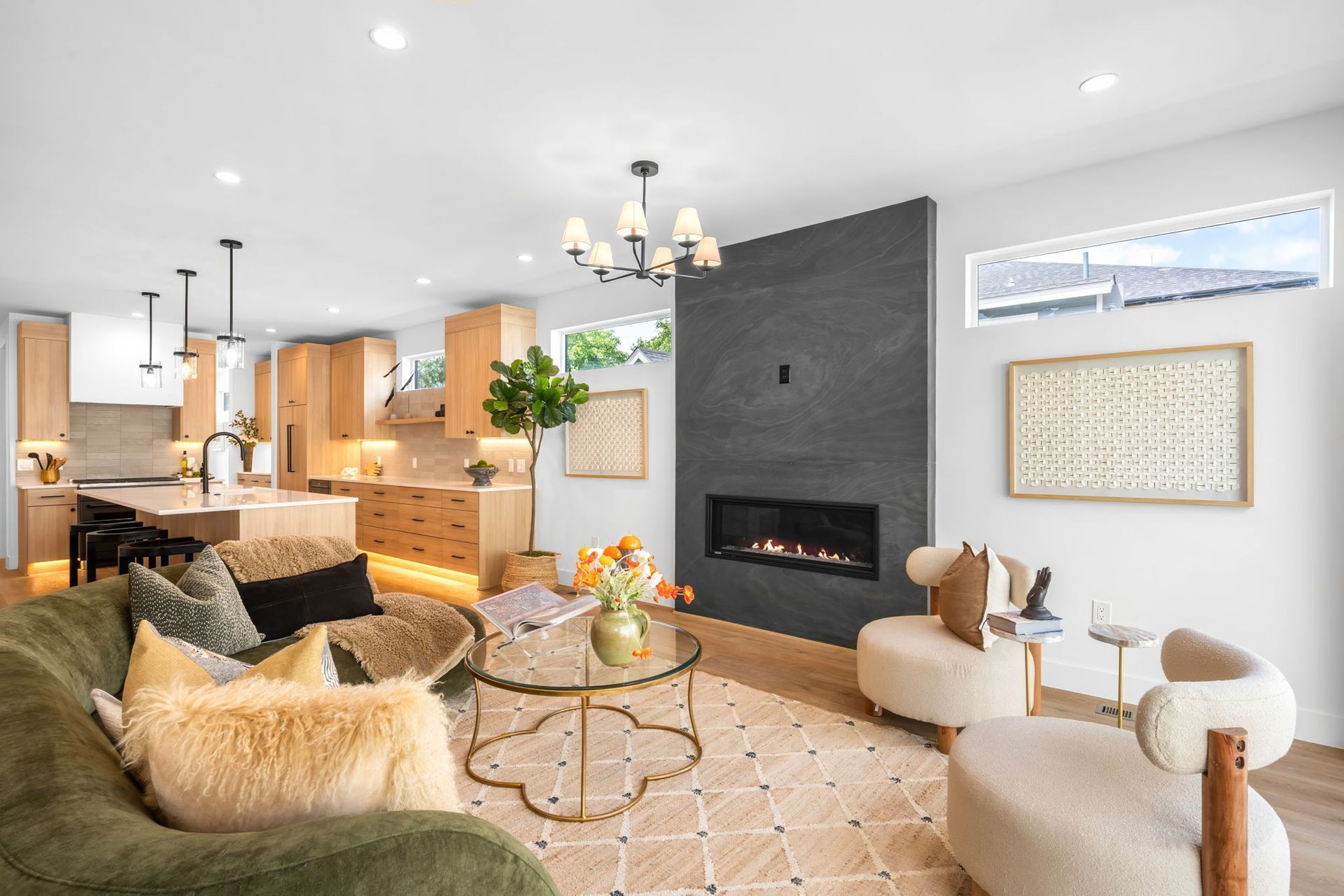 Modern living room with fireplace, kitchen, and light wood cabinetry. Olive green sofa, beige armchairs, and neutral rug.