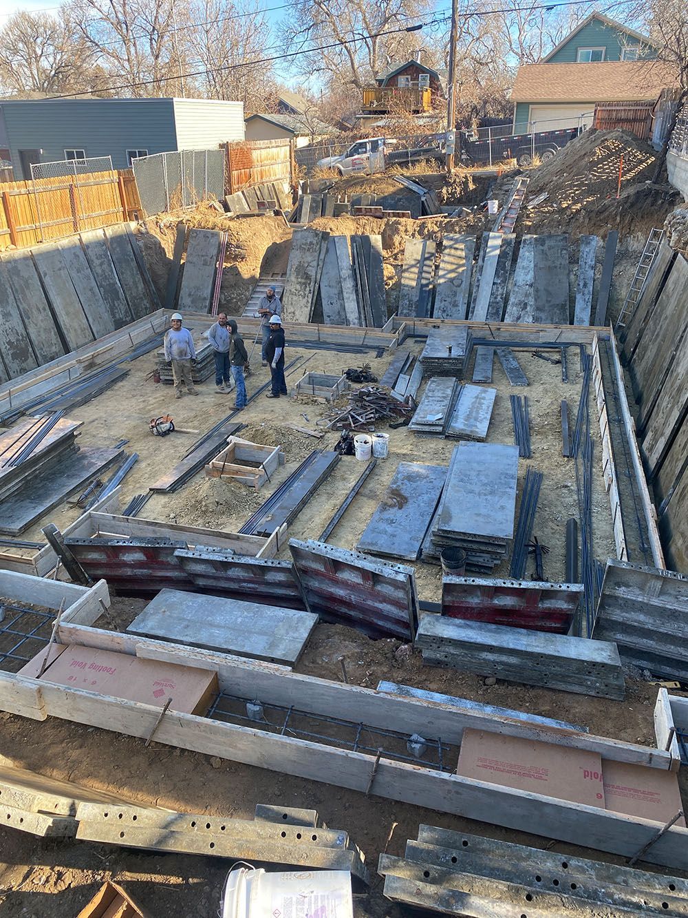 Construction site: workers inside a large excavated foundation pit, with concrete forms in place, preparing for concrete pouring.