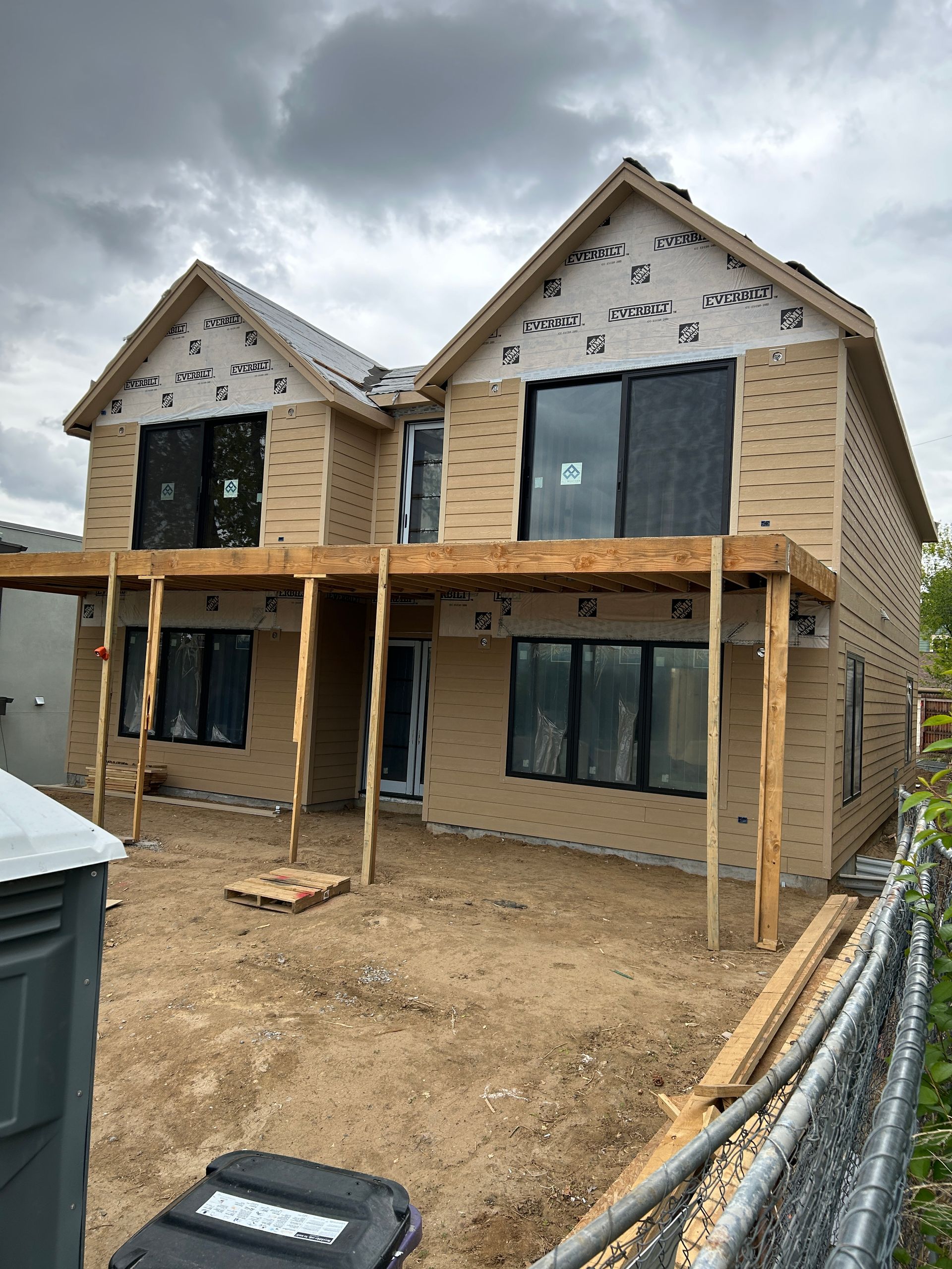 Two-story duplex under construction with wooden framing, tan siding, and black windows, under a cloudy sky.