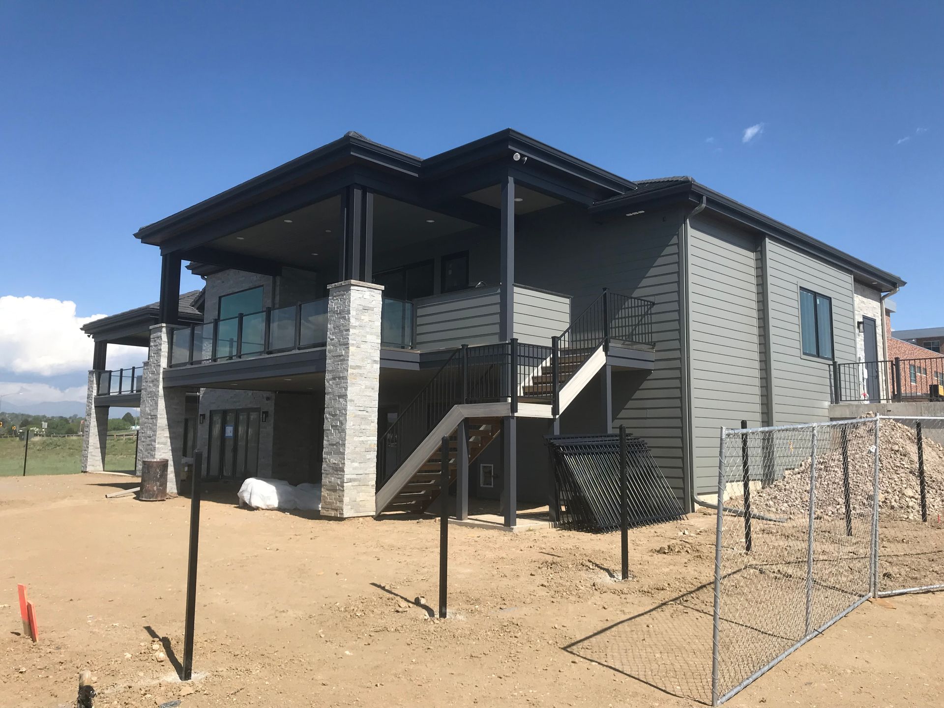 Two-story modern house under construction; gray siding, covered deck, stone pillars, blue sky.