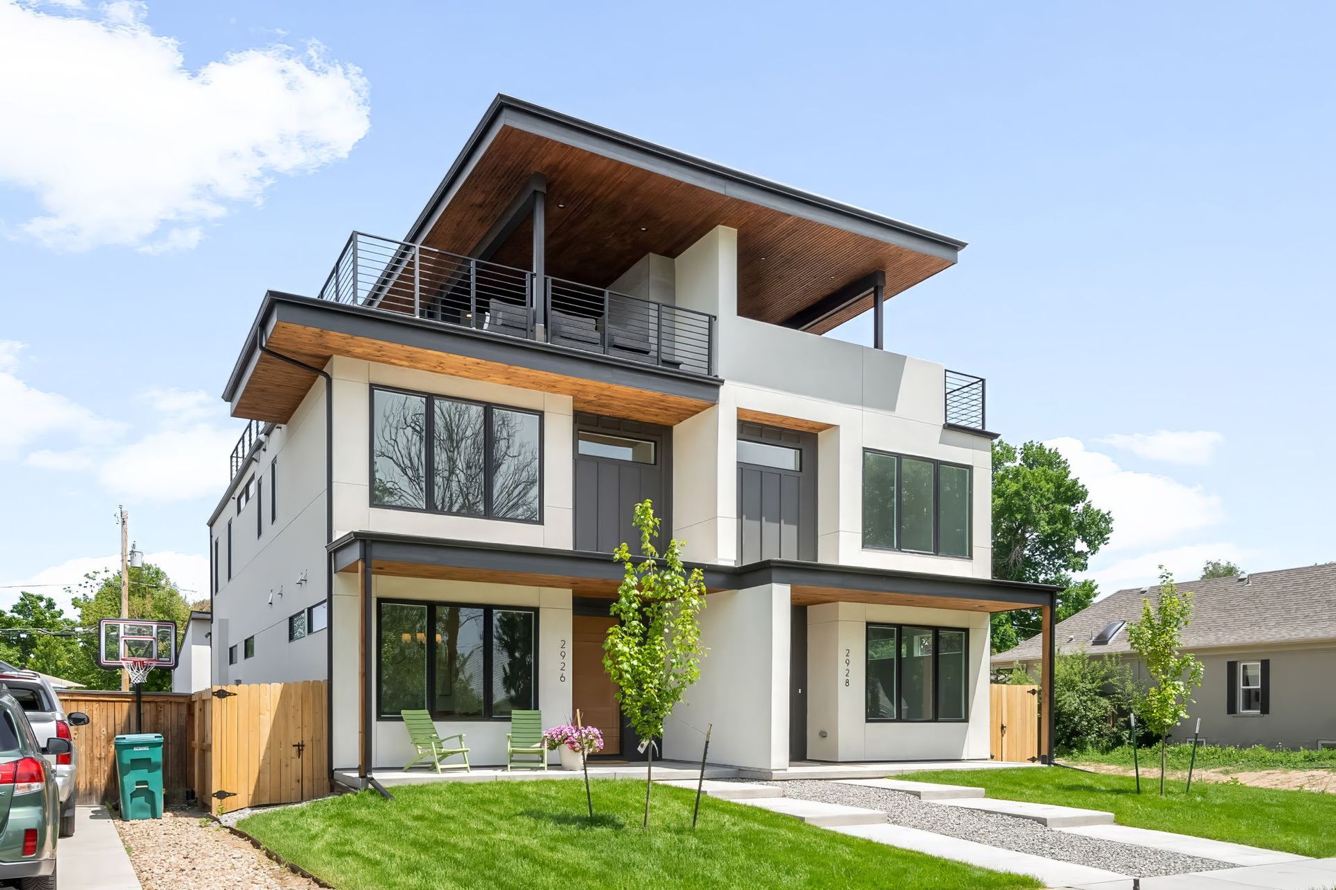 Modern three-story house with a flat roof, balcony, and wooden accents. Green lawn, blue sky, and a driveway.