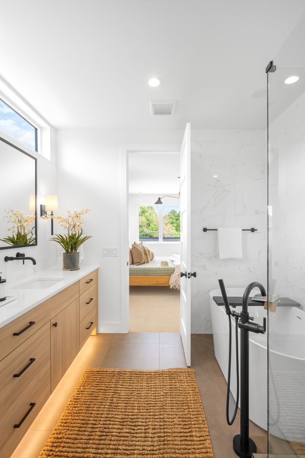 Modern bathroom with wood vanity, tub, and doorway to a bedroom. Neutral tones with a woven rug.