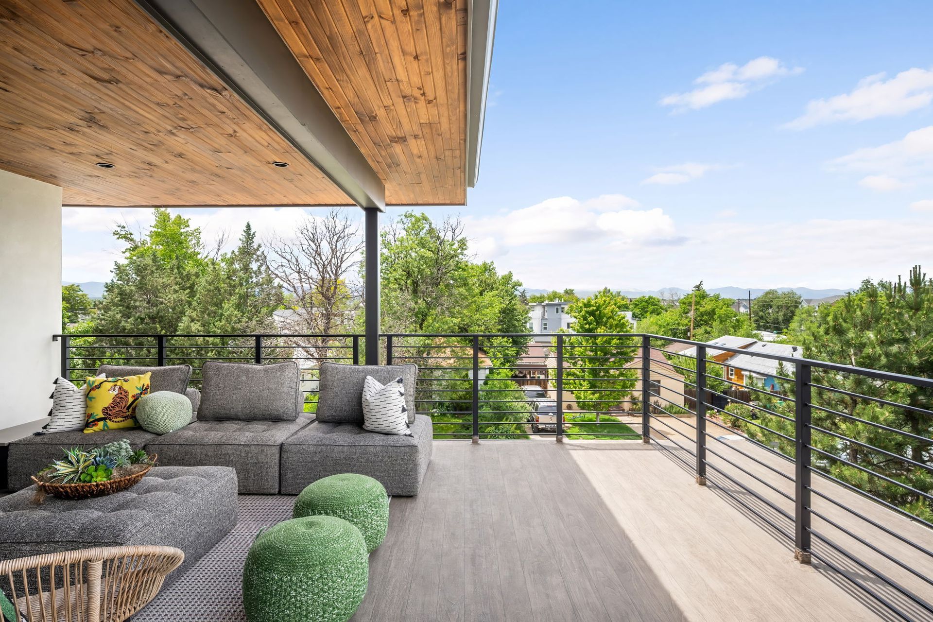 Outdoor patio with seating, wooden ceiling, and black railing overlooking trees and buildings on a sunny day.