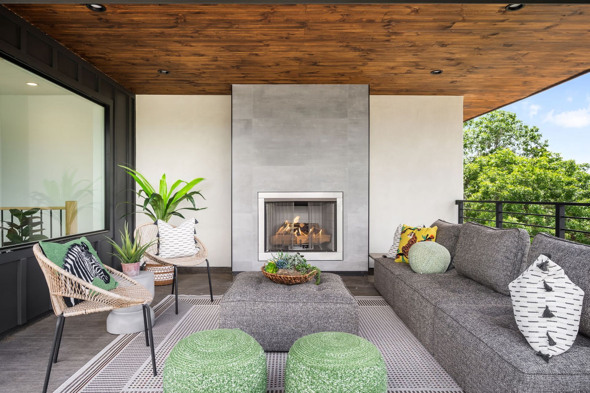 Outdoor living area with fireplace, gray sofa, woven chair, greenery, and wood ceiling.
