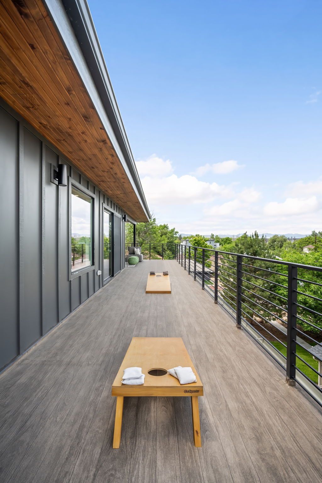 Long outdoor deck with cornhole boards and bean bags, overlooking trees and a blue sky.