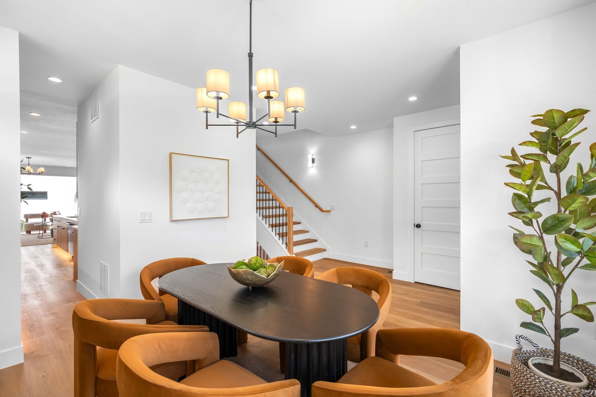 Dining room with oval black table, orange chairs, and chandelier; stairs in background.