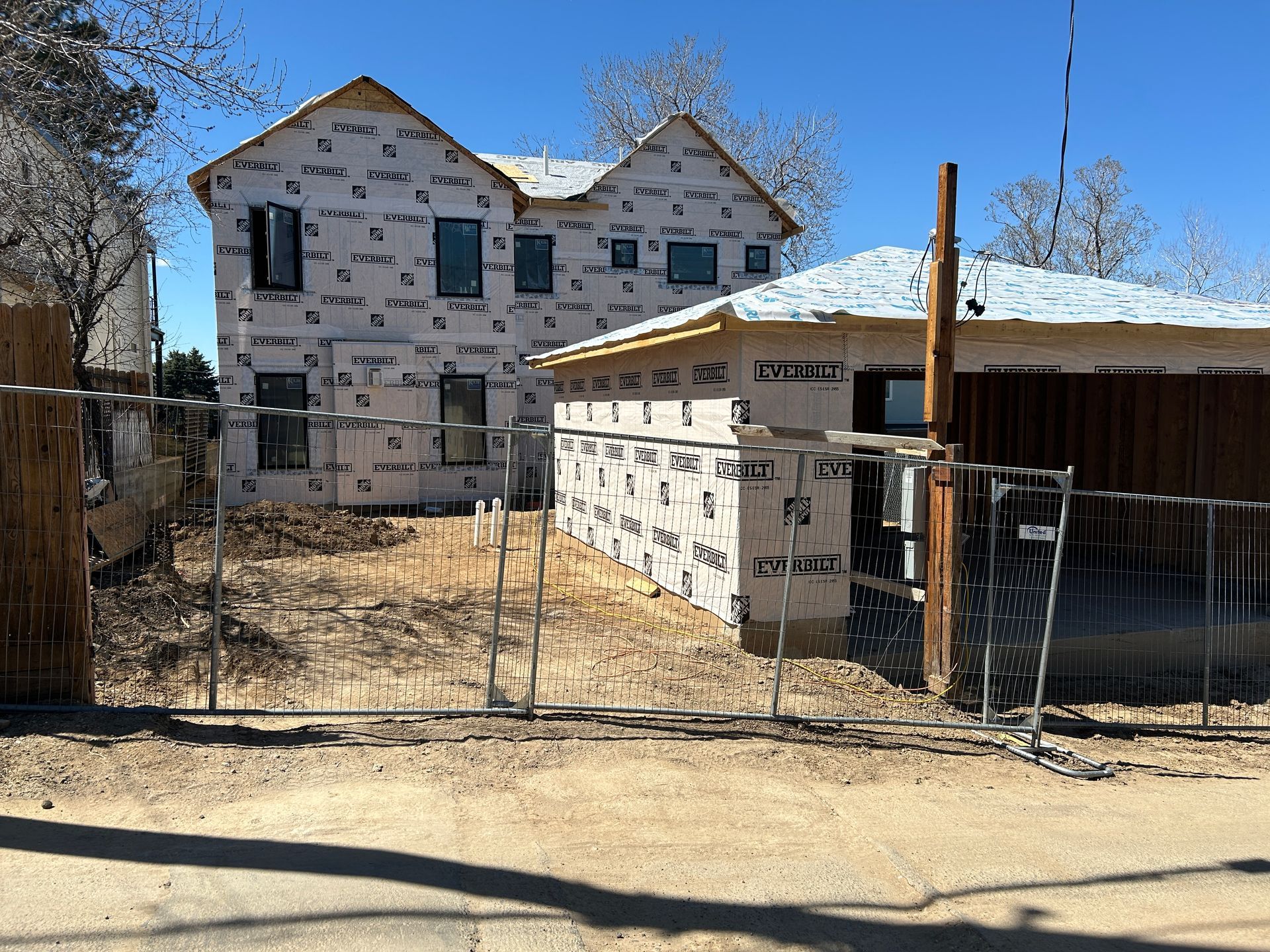 Two-story house under construction, wrapped in weather-resistant barrier, behind a chain-link fence on a sunny day.