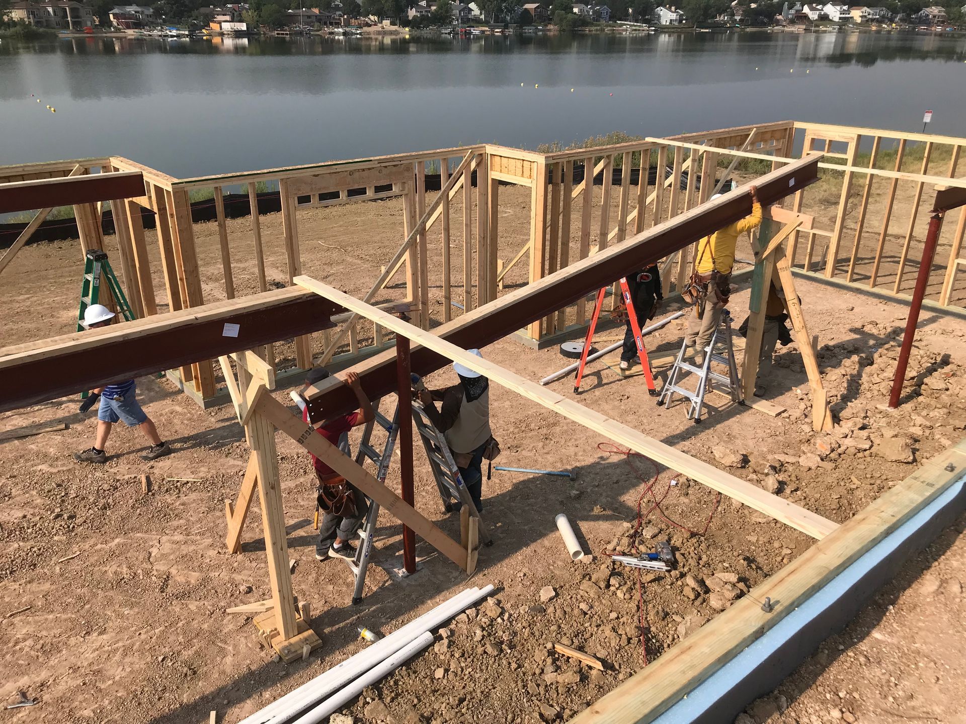 Construction site with wooden frame and metal beams; workers installing support structures. Lake in background.