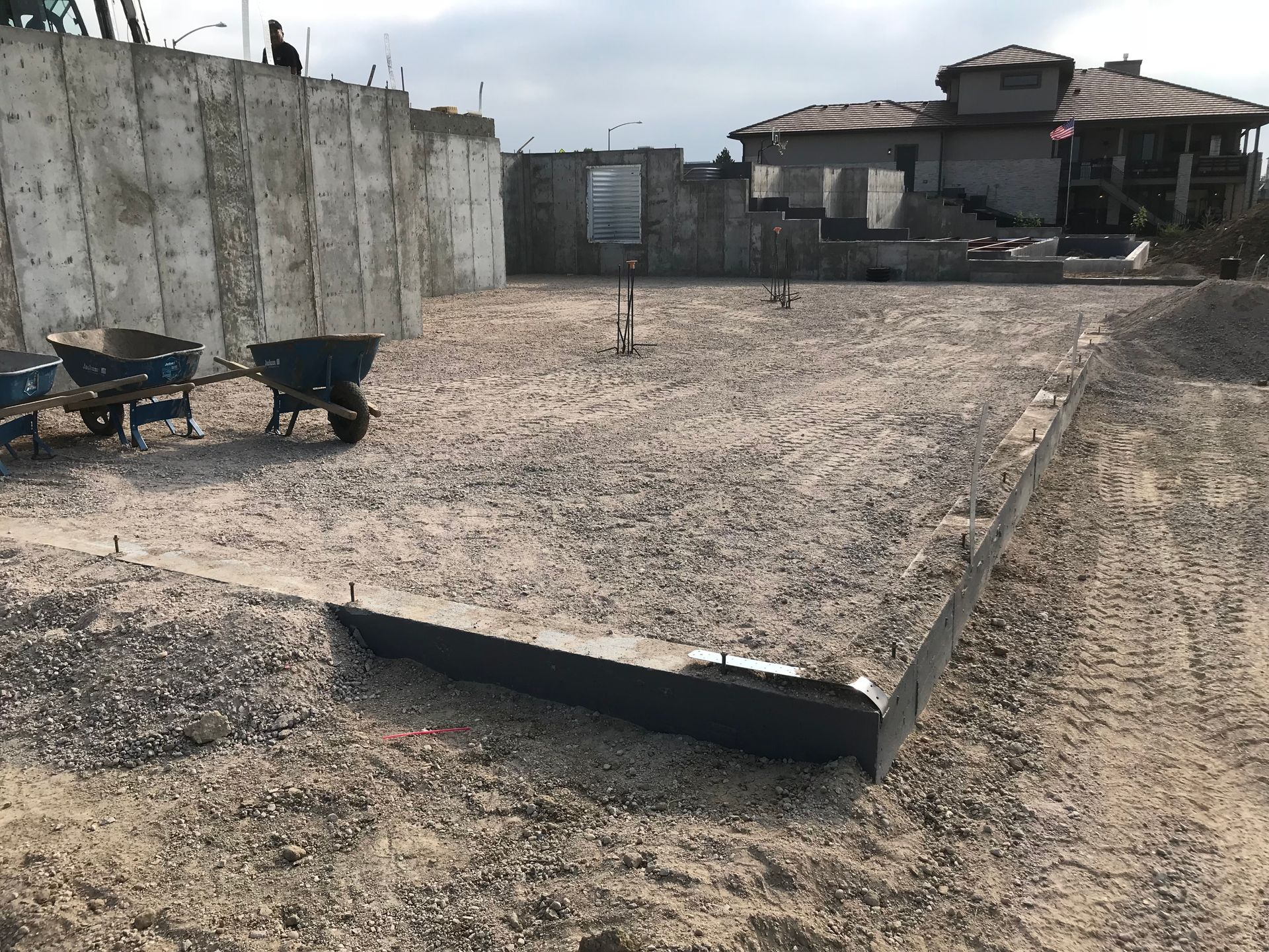 Construction site with gravel ground and concrete foundation.  Wheelbarrows sit near a large concrete wall; house in background.