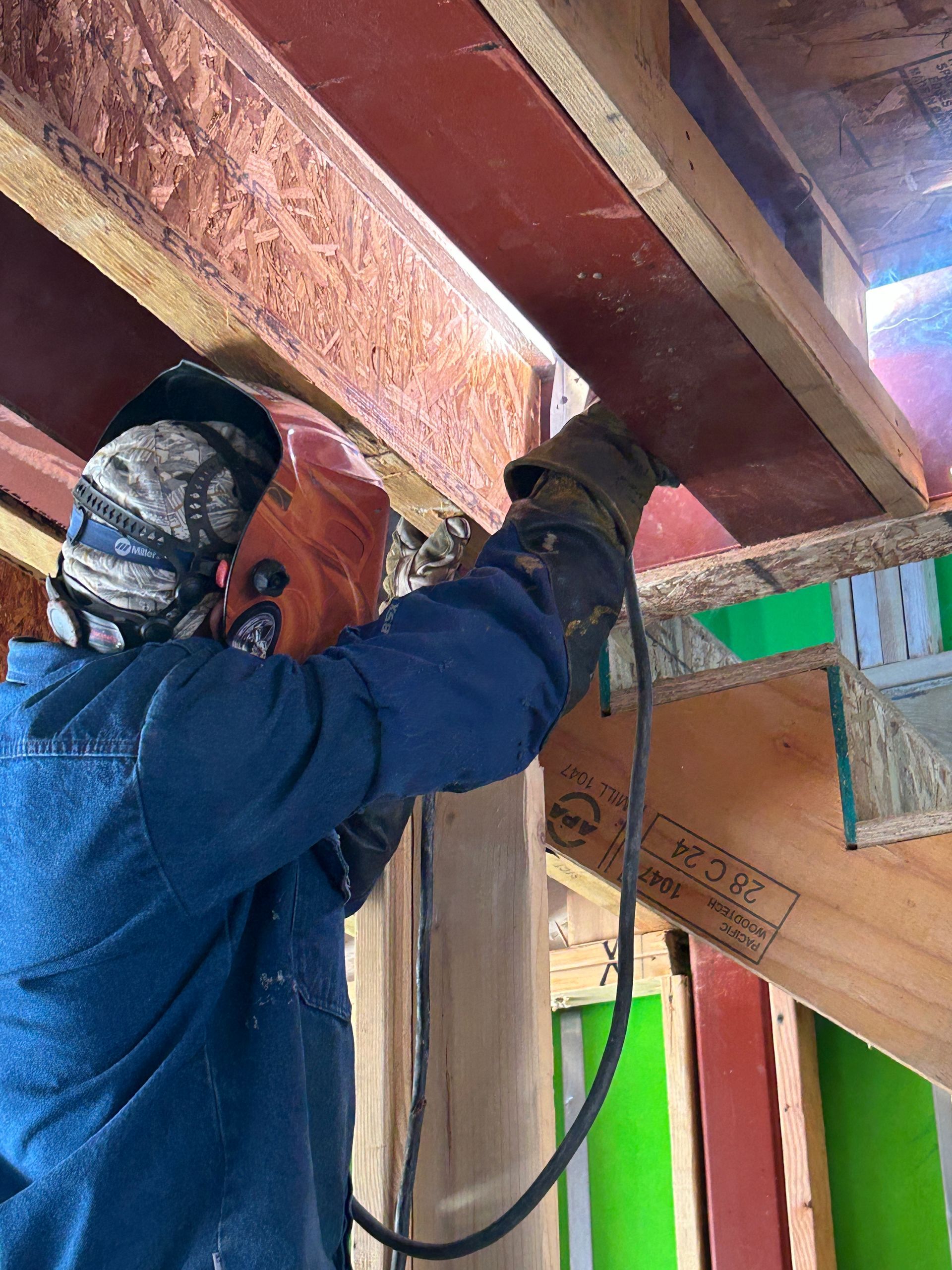 Person welding metal beams in a wood-framed structure. They wear a welding mask and gloves, using a welding torch.