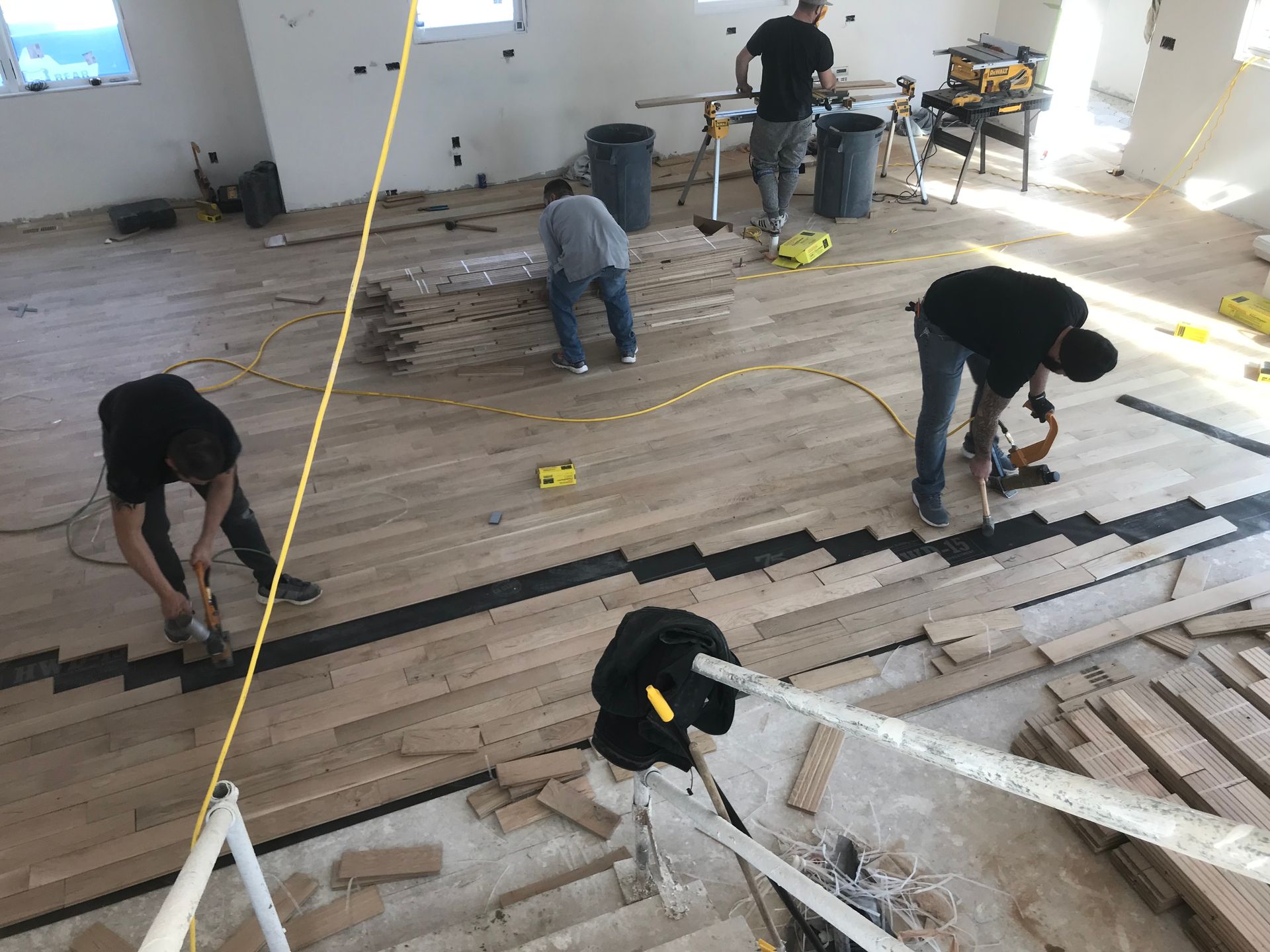Men installing hardwood flooring in a room. Boards are laid with black accents. Yellow cords and tools present.