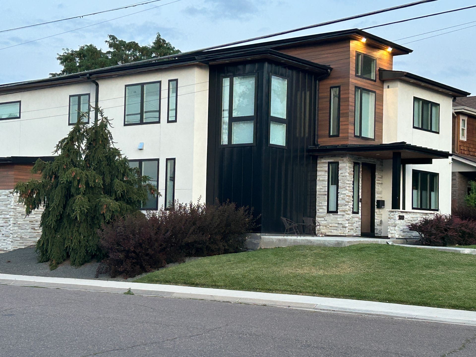 Modern two-story house with black trim, wood accents, white stucco, and stone facade. Green lawn and shrubs.