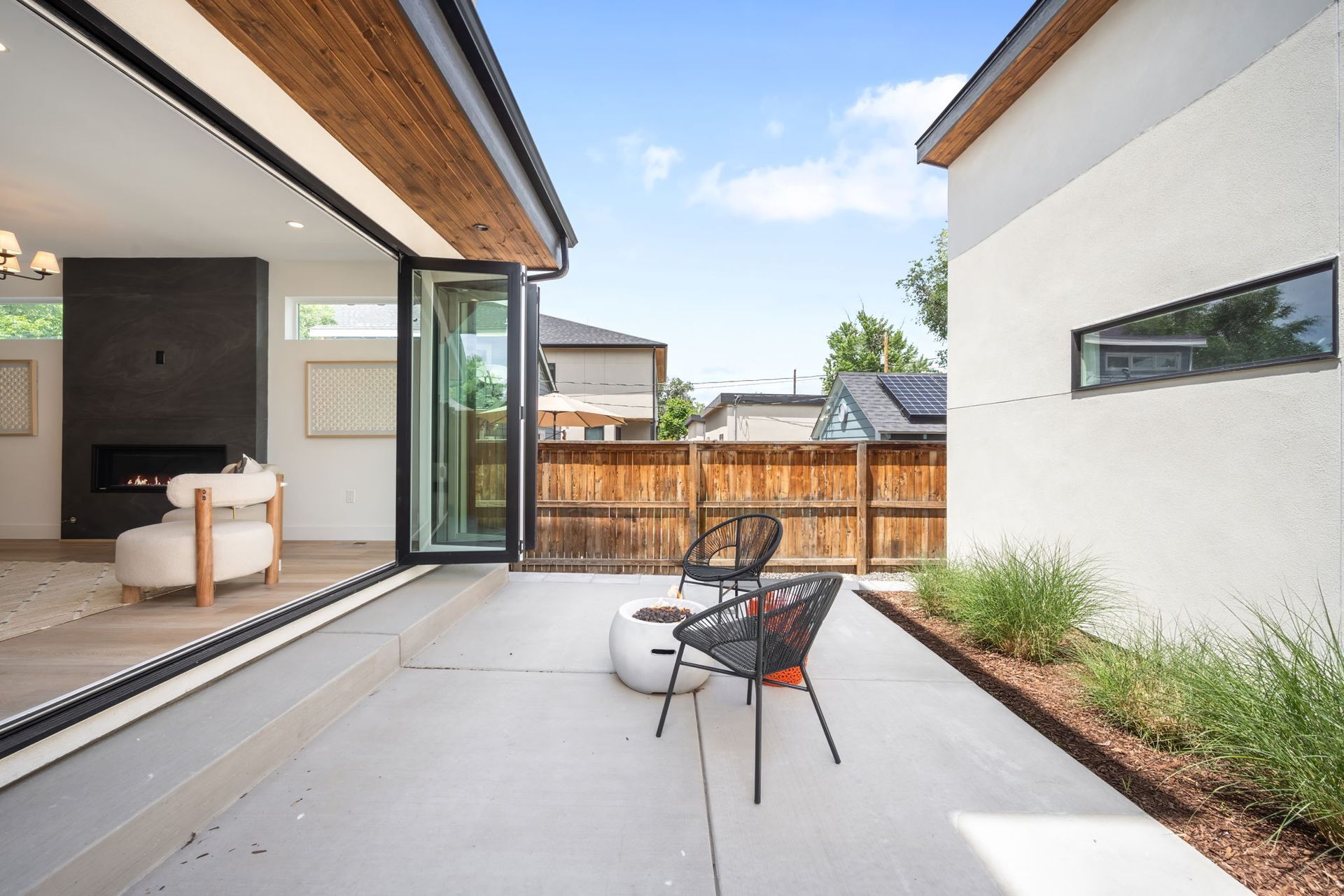Patio with modern chairs, fire pit, and open doors to a living room, against a wooden fence and blue sky.