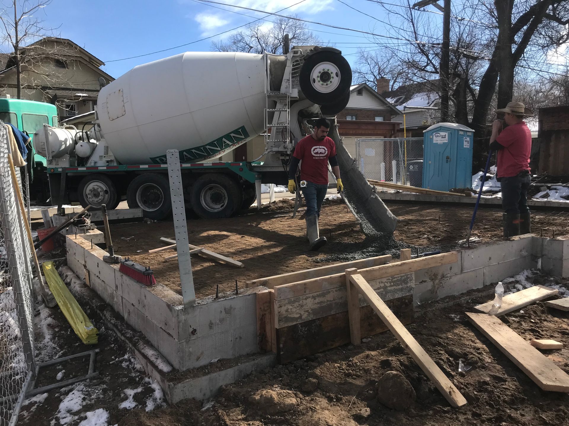 Concrete truck pouring concrete into a wooden form for a foundation; two workers supervise.