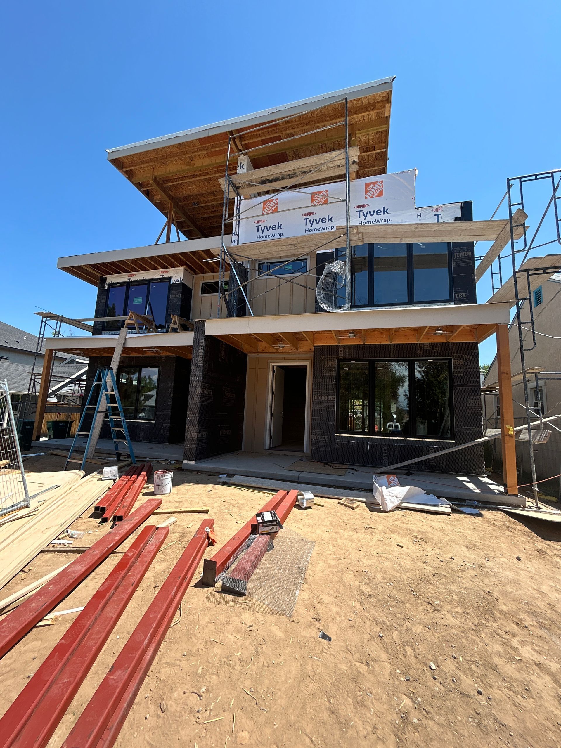 House under construction with stone facade, large windows, and exposed wood framing; blue sky.