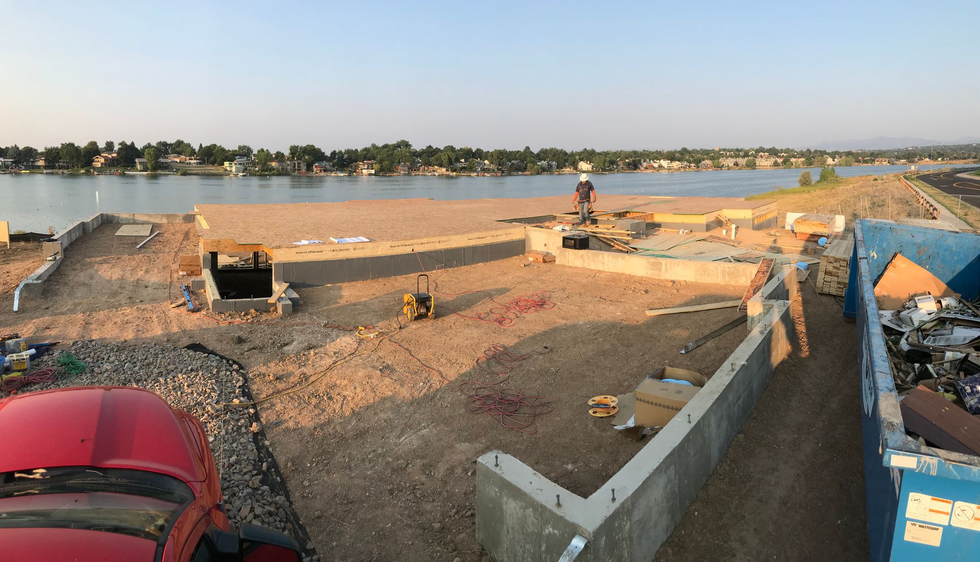 Construction site by a lake; red car, concrete structures, gravel, worker, blue trash bin, houses in the distance.