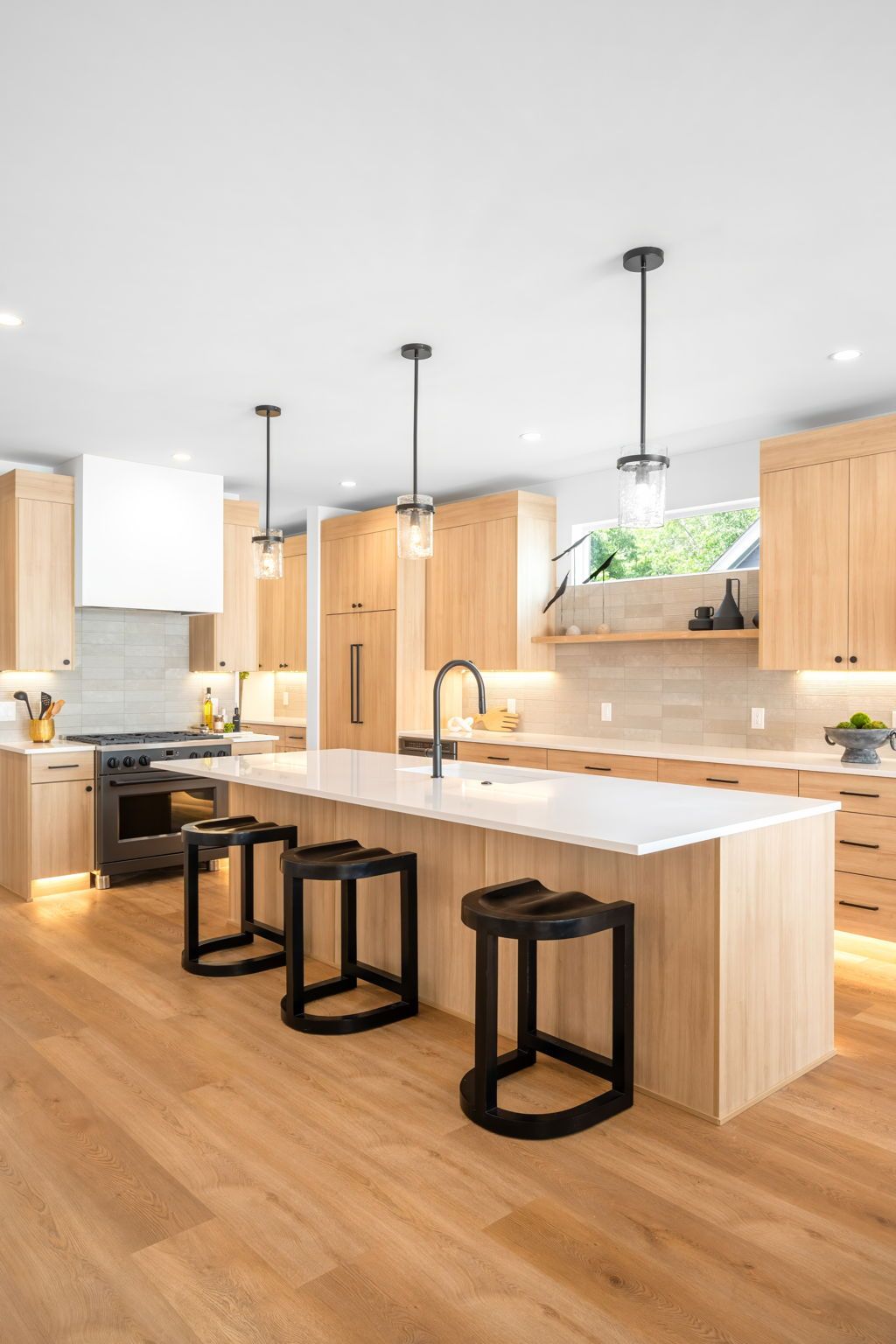 Modern kitchen with wood cabinets, white countertops, and black stools at the island.