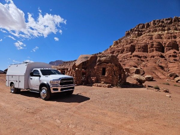 White truck with utility bed parked near a red rock structure under a blue sky.