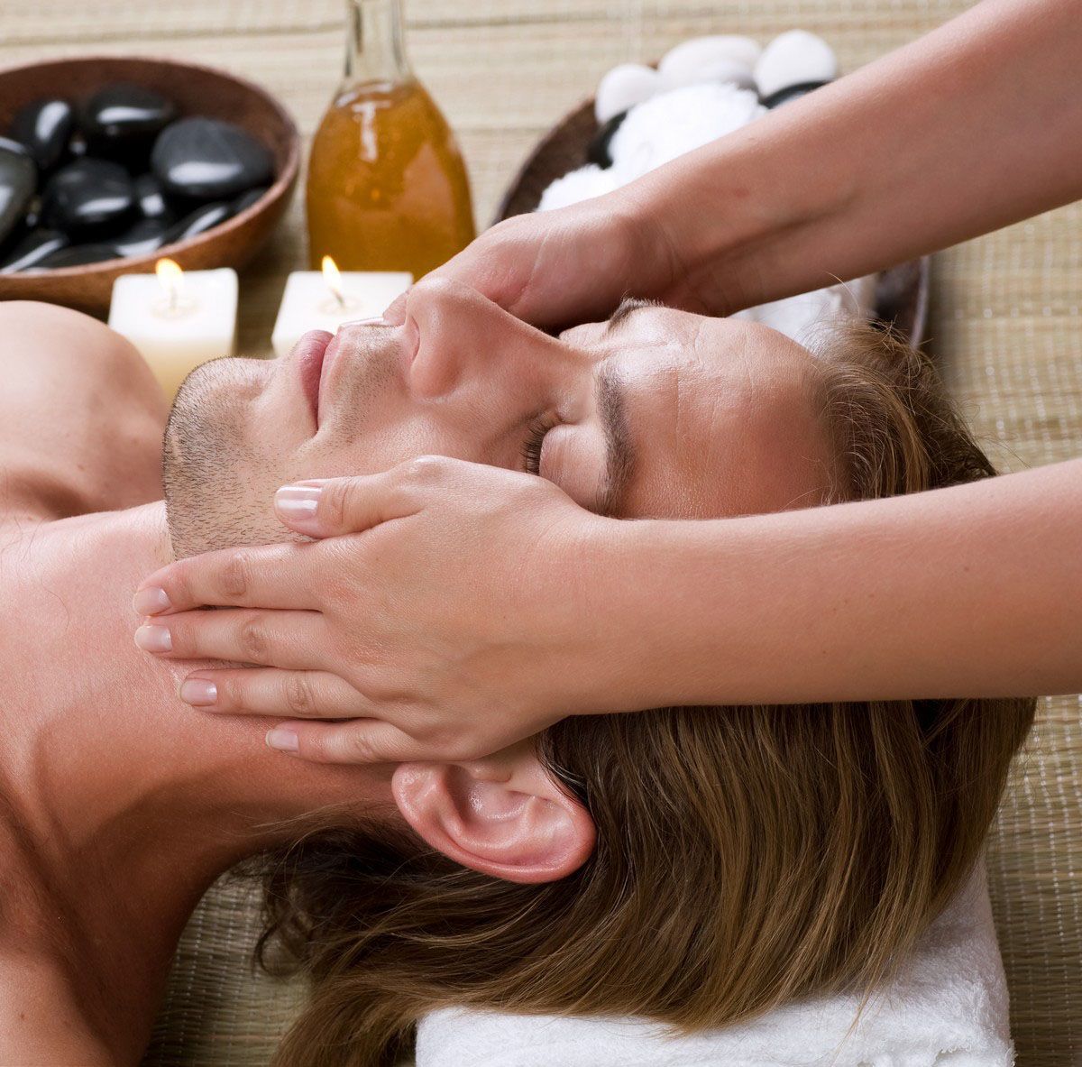 Man receiving a facial massage at a spa. Hands on face, candles, oil, and stones visible.