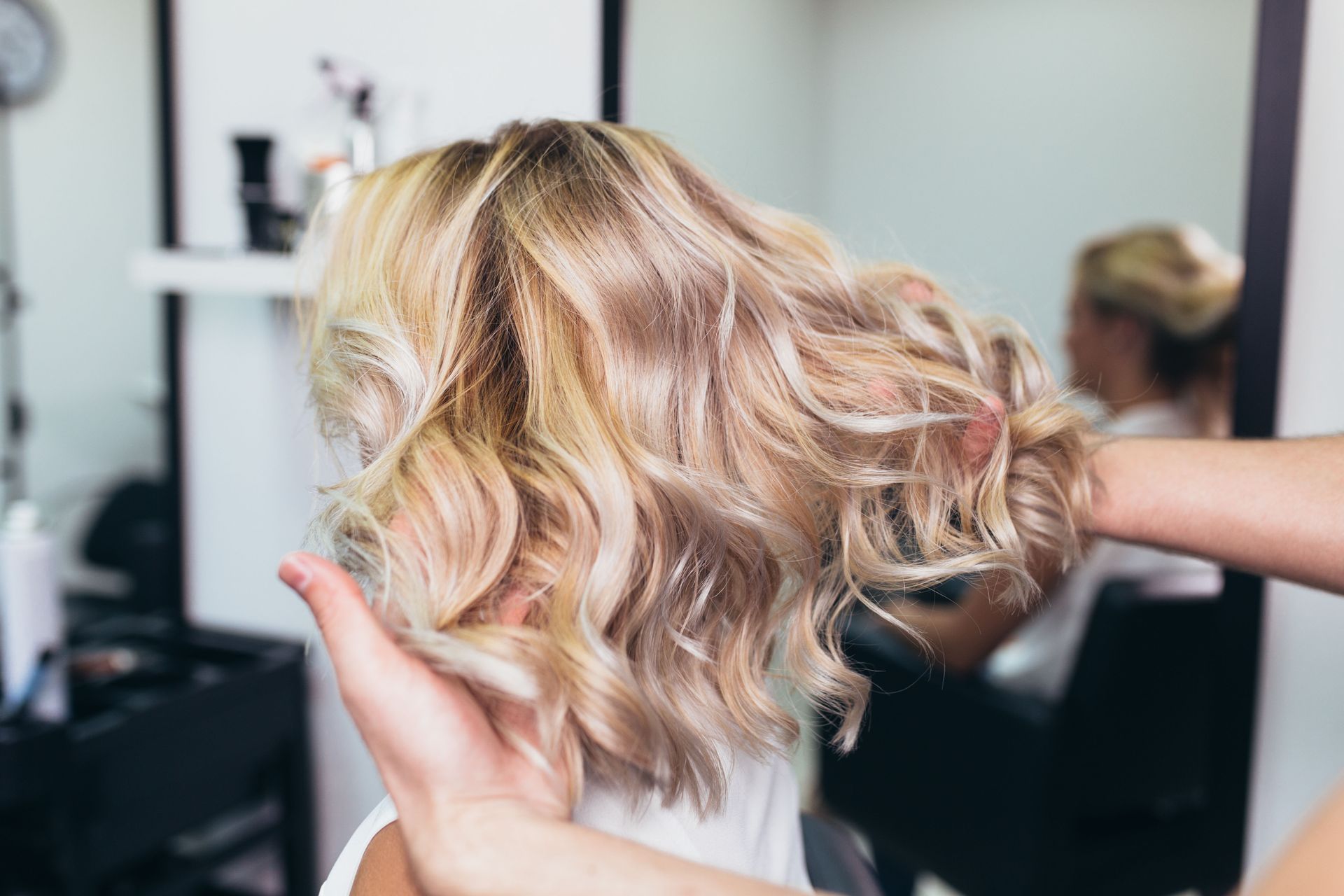 Person's styled blonde hair at a salon; stylist's hands hold the hair, mirror in background.