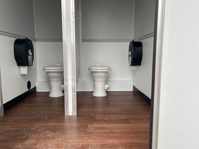 Restroom trailer with two toilets and a wooden floor.