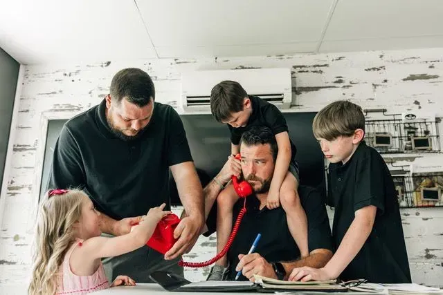 Man works at desk while four children interact with him and a red phone.