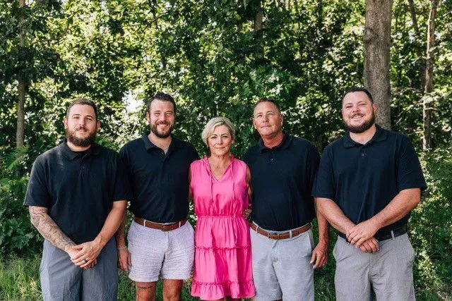Five people standing outdoors in front of trees; one woman in pink, others in black shirts and pants.