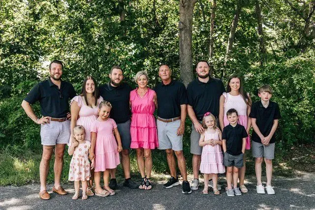 Large family posing outdoors; adults in navy shirts and shorts, children in pink.