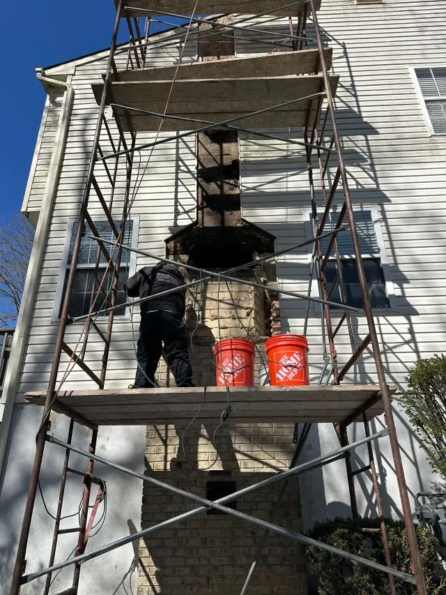 Person on scaffolding repairing chimney, two buckets on platform. Brick chimney against house.