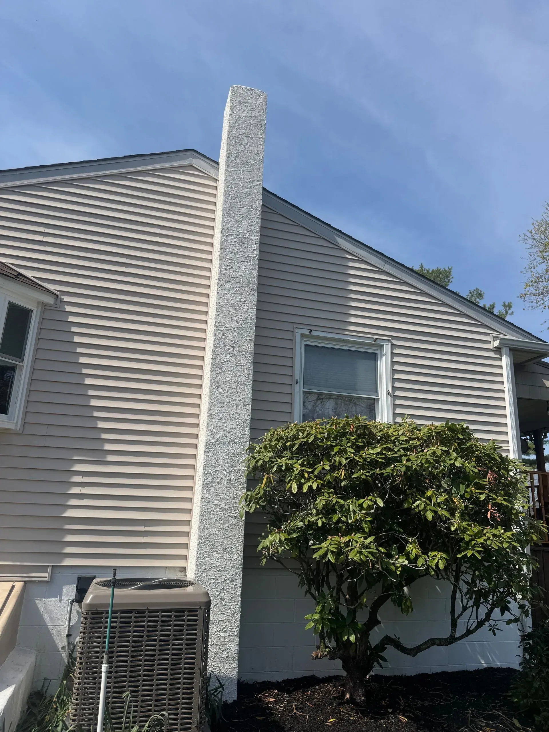 Chimney on side of a house with beige siding and a bush in front of a window.