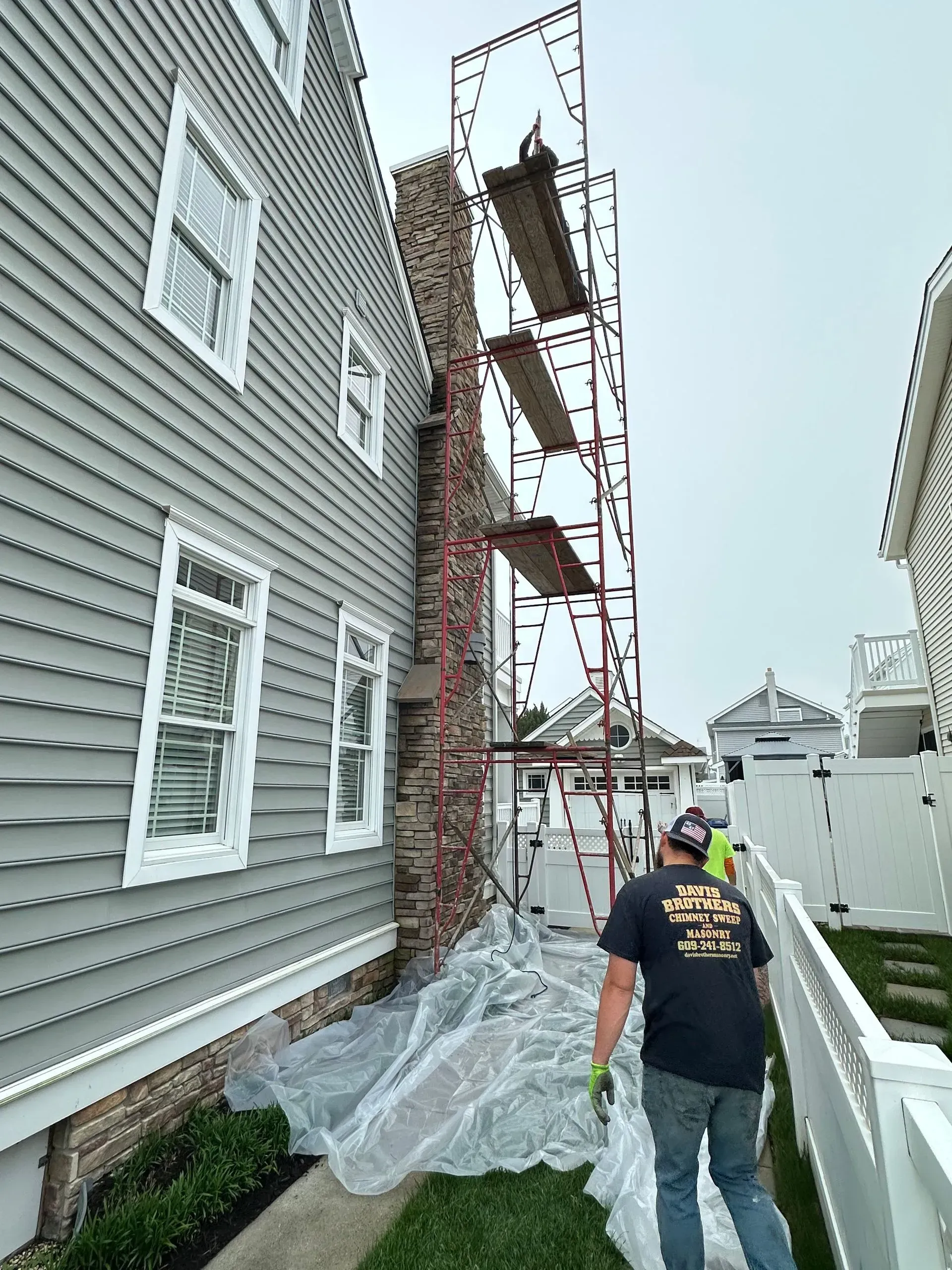 Chimney repair; scaffolding beside a brick chimney next to a gray house, worker in frame.