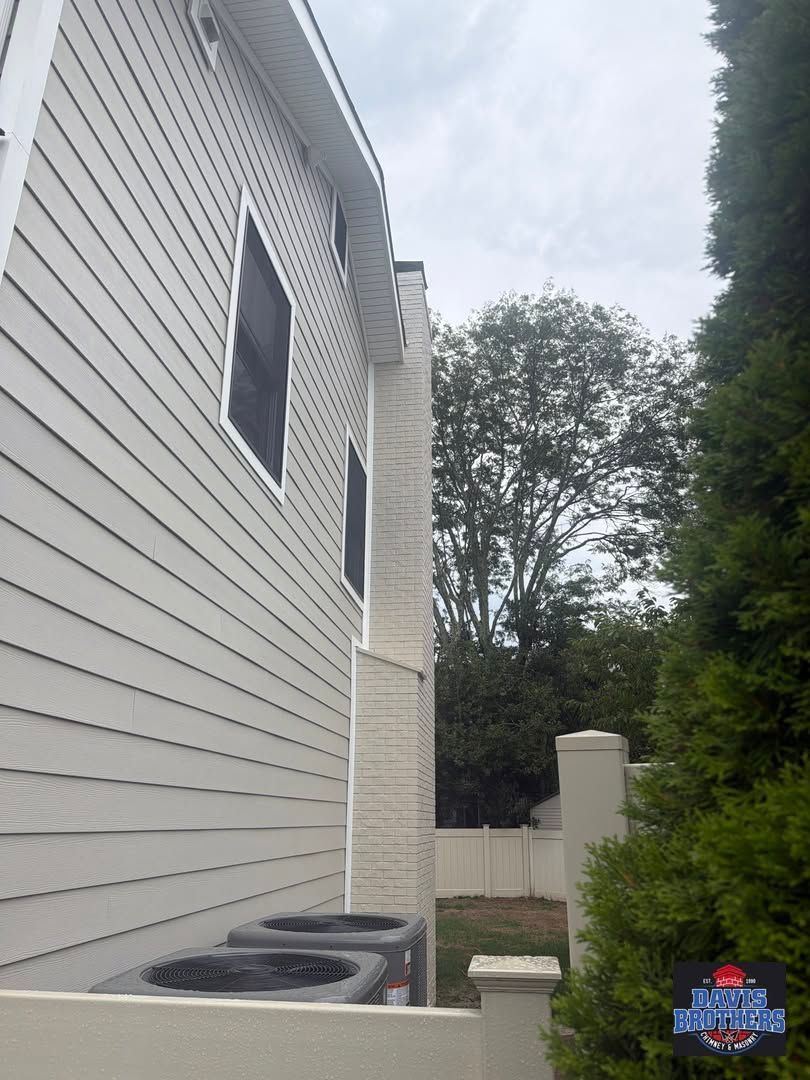 Side yard between a white house and tall evergreen, with air conditioner units and a fenced backyard.