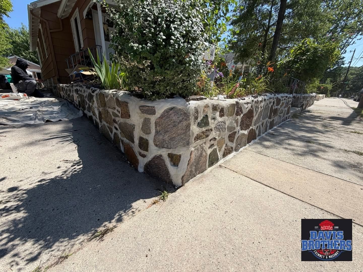 Stone planter wall with shrubs beside a sidewalk and building in sunlight