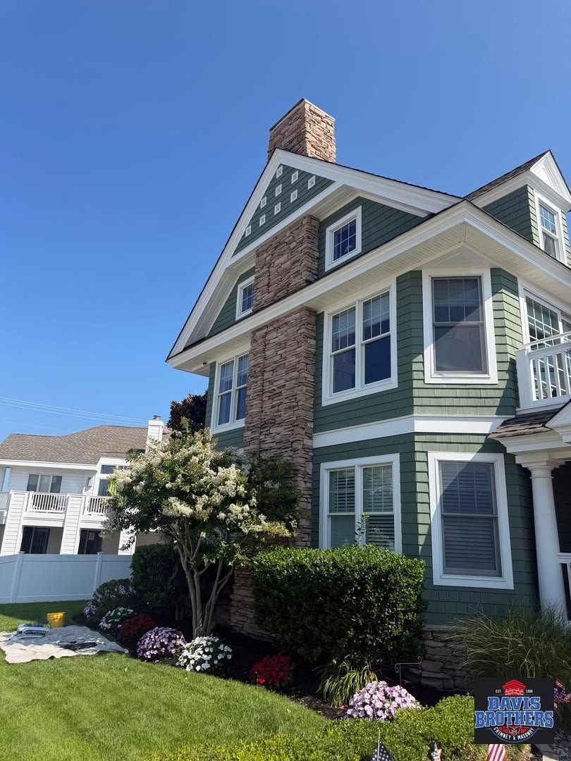 Green Victorian-style house with white trim, brick chimney, and flowered front landscaping under a blue sky