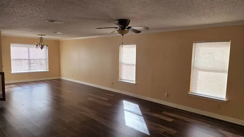 Empty living room with wood floors, tan walls, and three windows with shades. A ceiling fan and a light fixture are visible.
