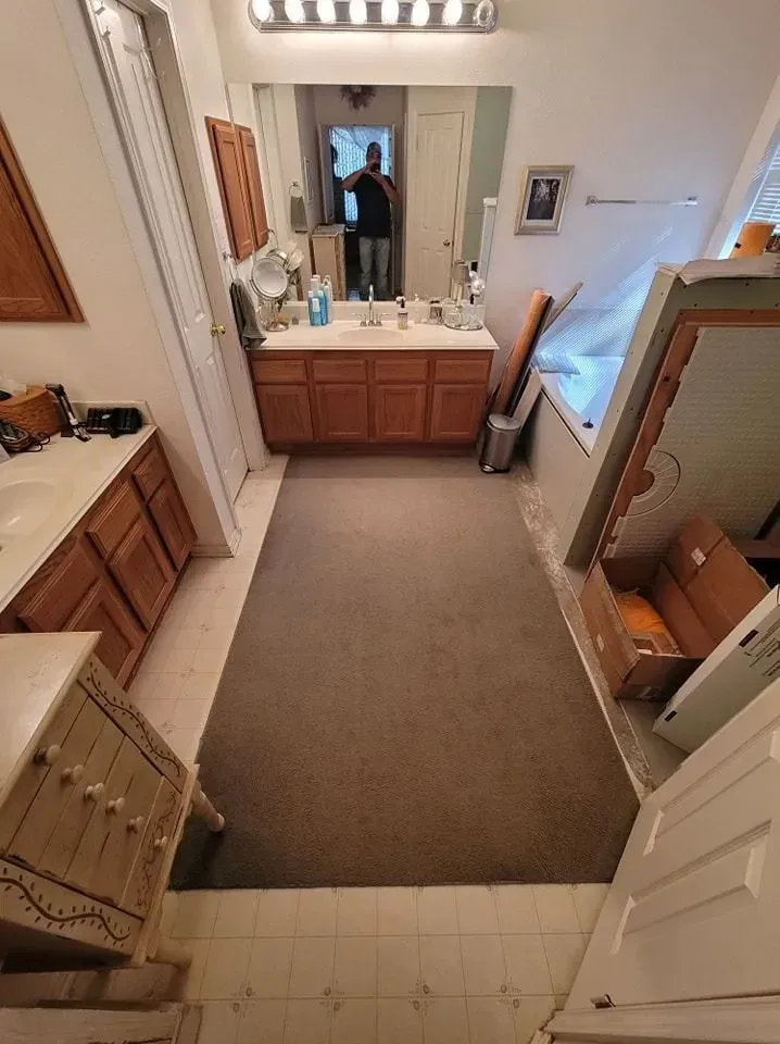 Bathroom with brown cabinets, mirror, tan carpet, and white walls. A person is reflected in the mirror.